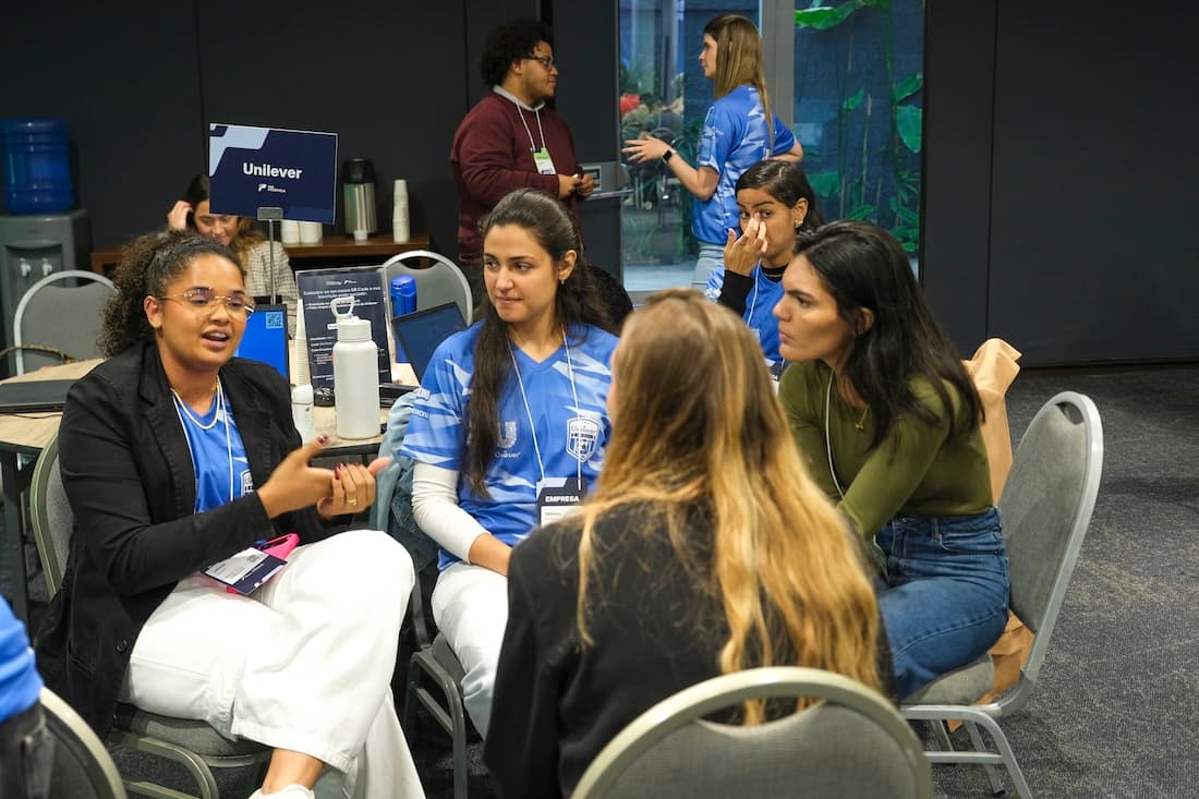 Três mulheres camiseta azul conversando com uma jovem em frente a uma placa da Unilever, onde o trio trabalha.